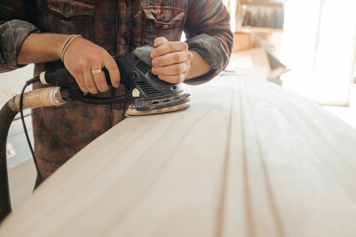 Hands guiding an orbital sander with dust extraction across a flat panel.
