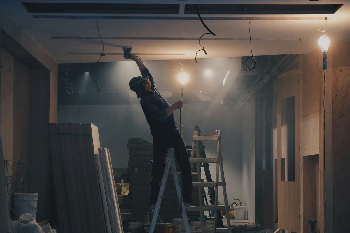 Worker on a stepladder reaching up to work on a ceiling in a room under renovation.