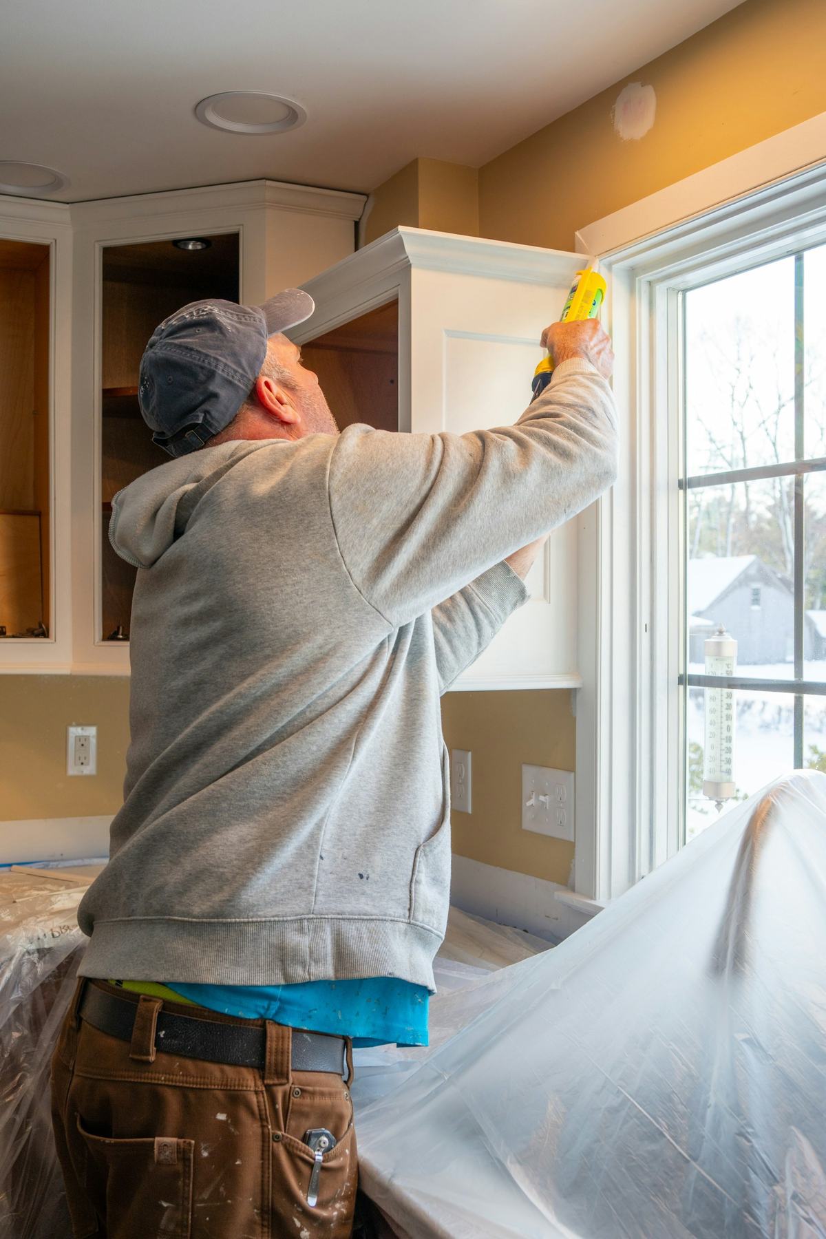 Painter seen from behind caulking a window casing, with plastic sheeting protecting the surfaces below.